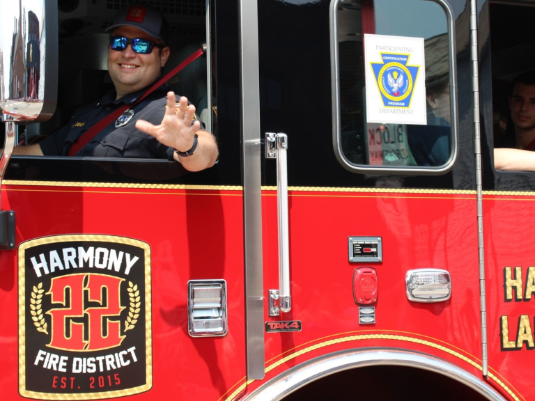 A firefighter in uniform smiles and waves from the driver's seat of a red fire truck labeled "Harmony Fire District 22." Another person is partially visible in the passenger seat.