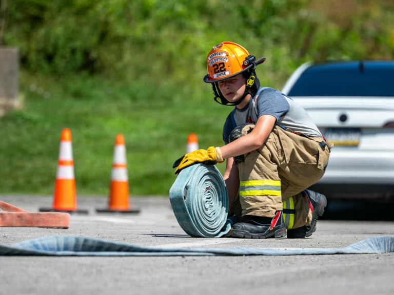 A firefighter in uniform kneels on pavement, rolling up a hose. Traffic cones and a white car are in the background on a sunny day.