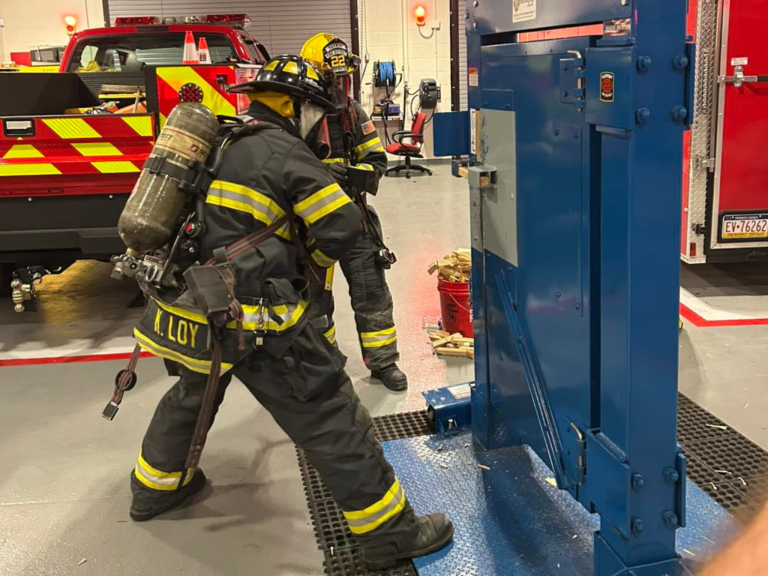 Two firefighters in full gear use heavy equipment to practice forcible entry during a training exercise in a fire station. Fire trucks and tools are visible in the background.