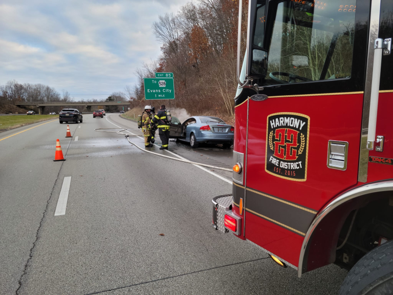 Firefighters stand next to a silver car by the roadside, with fire hoses on the ground. A fire truck marked “Harmony Fire District” is in the foreground. Traffic cones partially block the road under a cloudy sky.