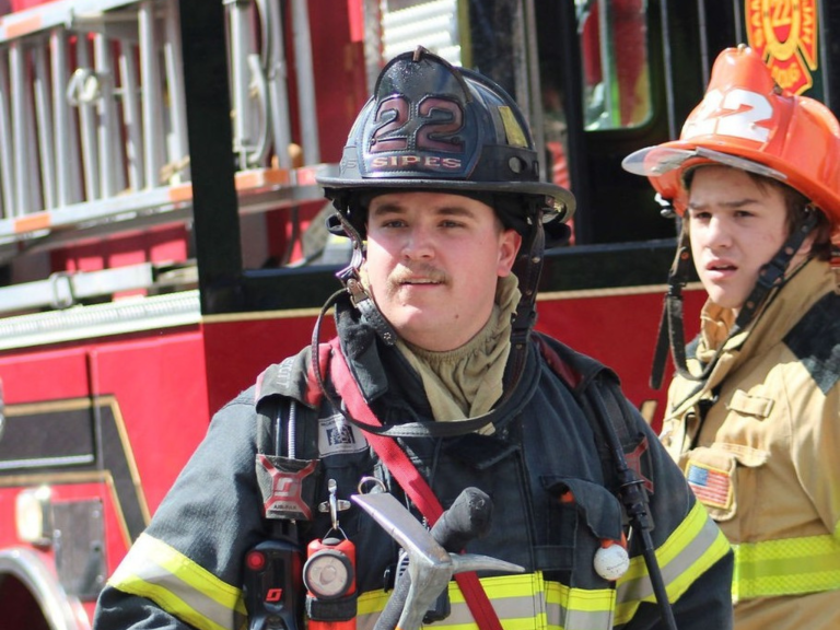 Two firefighters in full gear, including helmets and jackets, stand in front of a red fire truck. One firefighter is in the foreground holding a tool, while the other stands slightly behind.